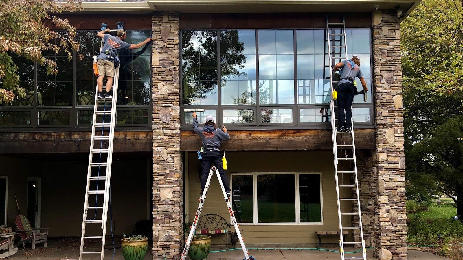 Three Walton's crew members on ladders washing the large windows of a two-story stone-and-wood home.