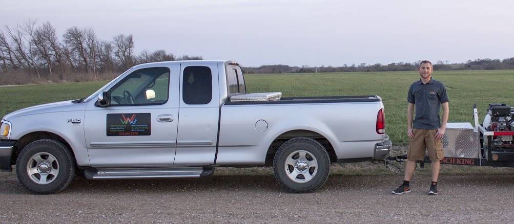 A silver pickup with a services and phone-number sign on the door, parked on a gravel road beside a field, with a pressure washer trailer and a crew member standing at the right.